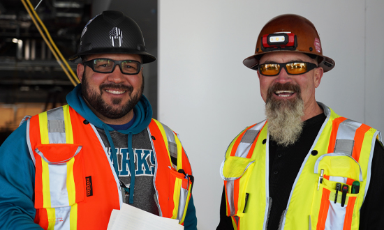 Two men with beards wearing construction hats and vests, one holding papers, smiling to the camera.