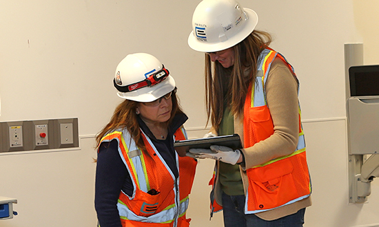 Two women in construction protective gear, one holding an iPad and both looking at it in discussion.