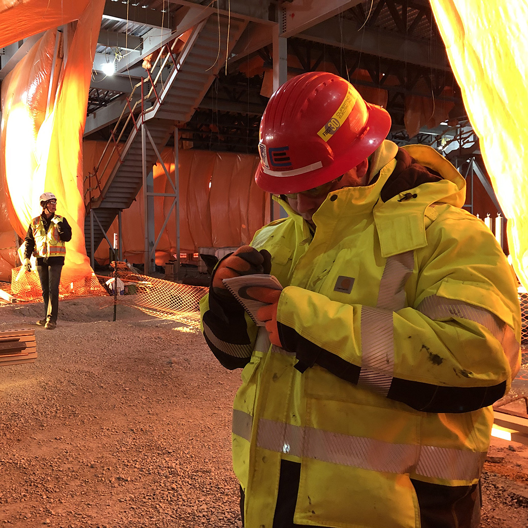 A man wears a yellow safety jacket and red hard hat and writes in a small notebook.