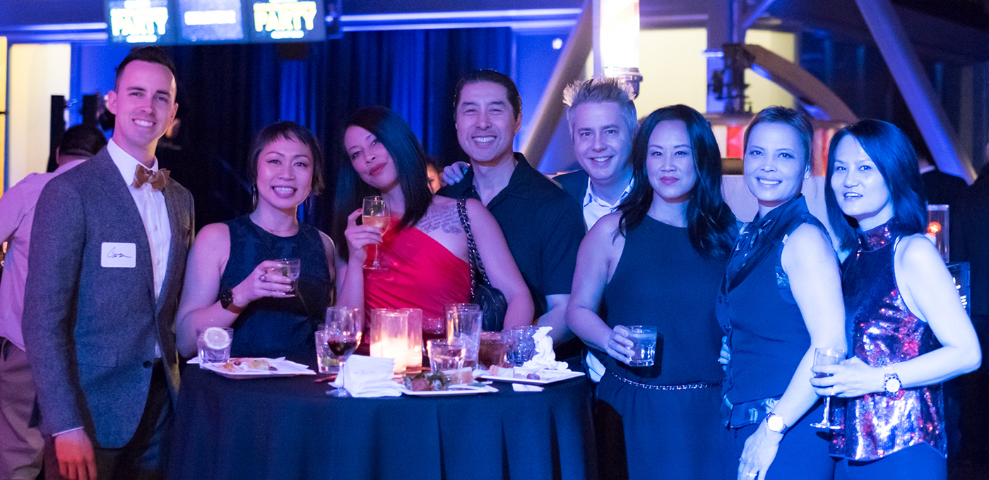 Five women and four men dressed in cocktail attire at a party, some holding cocktails, all smiling for the camera.