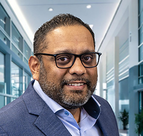 Man with light brown skin named Janesh with black rimmed glasses smiling at the viewer, wearing a light blue shirt and muted blue jacket.