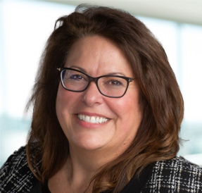 Light skinned woman with medium brown hair in a black jacket wearing black-rimmed glasses, smiling with teeth showing